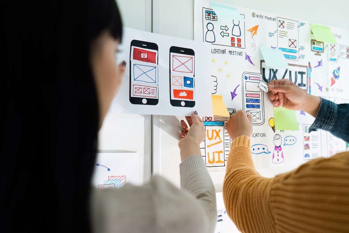 Des personnes placent des schémas de design UX et SEO colorés sur un tableau blanc, illustrant des interfaces mobiles et des notes adhésives.
